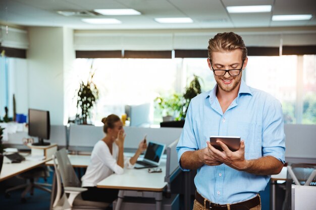 Young successful businessman smiling, looking at tablet, over office