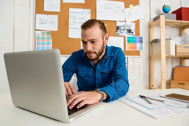 Free photo young successful businessman smiling, sitting at workplace typing on laptop