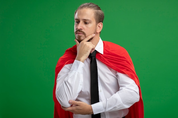 Free photo young superhero guy putting hand on chin isolated on green