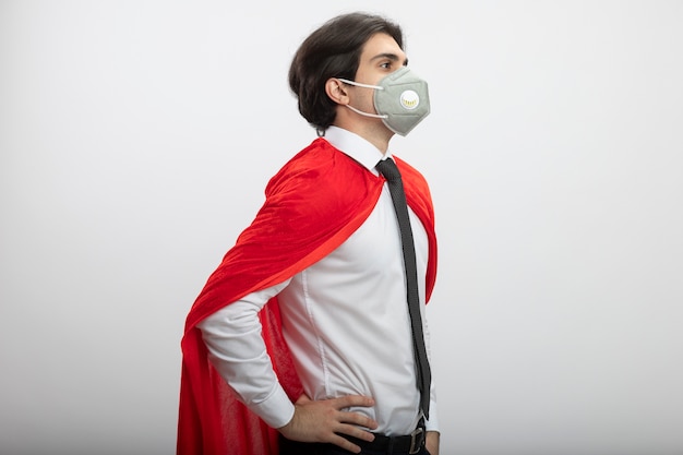 Free photo young superhero guy standing in profile view wearing tie and medical mask putting hands on hip