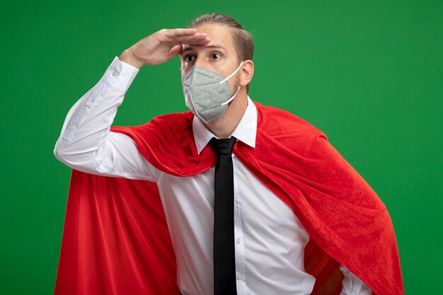 Young superhero guy wearing medical mask and tie looking at distance with hand isolated on green