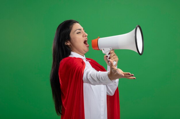 Young superwoman standing in profile view talking in loud speaker looking straight stretching out hand isolated on green wall