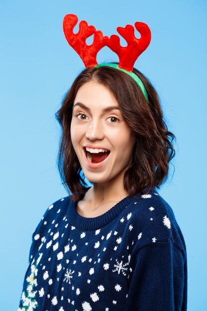 Young surprised beautiful brunette girl in knitted sweater and christmas reindeer antlers smiling  over blue wall