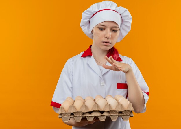 Young surprised blonde female chef in chef uniform holds and looks at batch of eggs isolated on orange wall