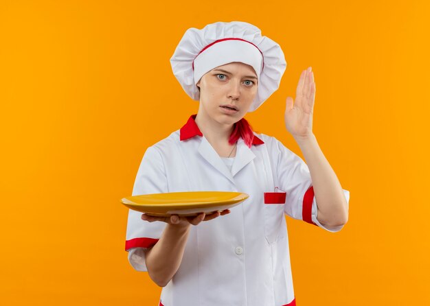 Young surprised blonde female chef in chef uniform holds plate and raises hand up isolated on orange wall