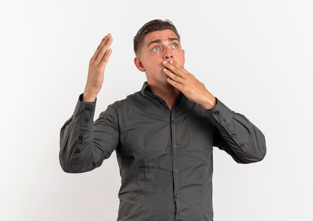 Young surprised blonde handsome man puts hand on mouth and raises hand up isolated on white background with copy space