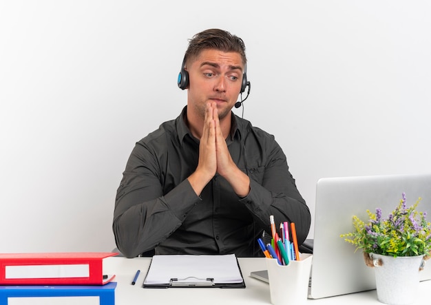 Young surprised blonde office worker man on headphones sits at desk with office tools looking at laptop holds hands together isolated on white background with copy space