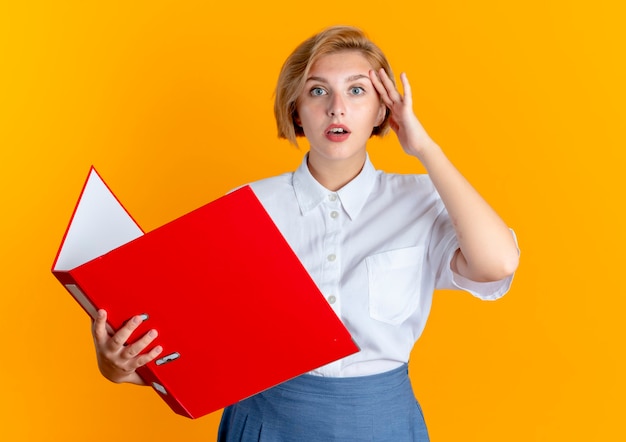 Free photo young surprised blonde russian girl puts hand on head holding file folder isolated on orange background with copy space