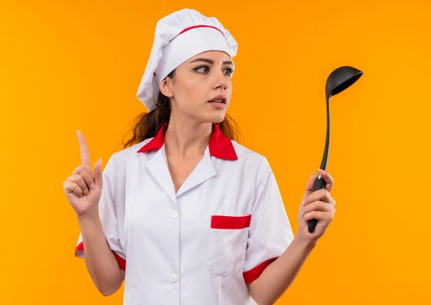 Young surprised caucasian cook girl in chef uniform holds ladle and points up isolated on orange wall with copy space