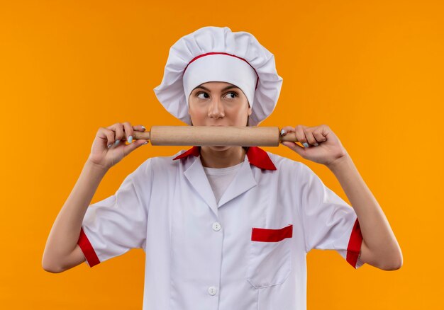 Young surprised caucasian cook girl in chef uniform holds and pretends to bite rolling pin isolated on orange background with copy space