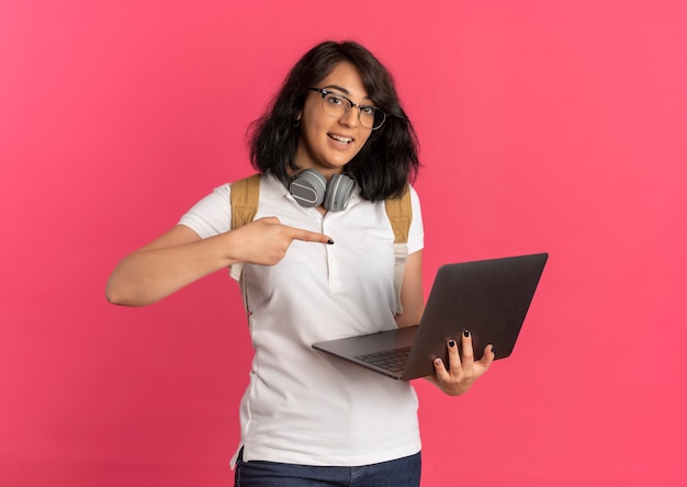 Young surprised pretty caucasian schoolgirl with headphones on neck wearing glasses and back bag holds and points at laptop on pink  with copy space