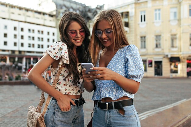 Young tanned girls in denim pants and stylish blouses look at purple phone screen Blonde and brunette women in colorful sunglasses smile and talk outside