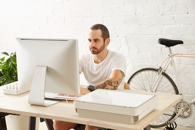 Free photo young tattooed freelancer man in blank white t-shirt works on his computer at home near his bicycle, looking in display