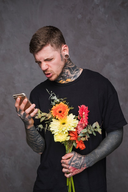 Young tattooed and pierced man holding flower bouquet in hand making face while using mobile phone