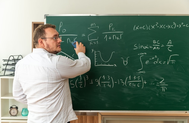 young teacher wearing glasses standing in behind view in front of chalkboard in classroom looking at side cleaning chalkboard with chalk eraser