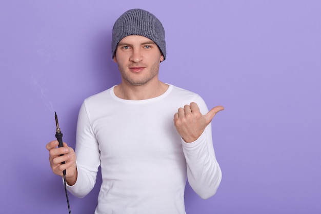 Young technician being ready to solder wire, attractive male wears white casual shirt and gray cap holds soldering iron in one hand and point aside with another thumb, isolated on purple.