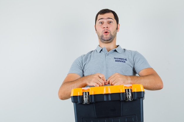 Free photo young technician in grey uniform holding toolbox with his both hands and looking surprised.