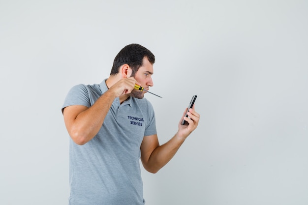 Young technician holding screwdriver and trying to open the back of his phone in grey uniform and looking focused.