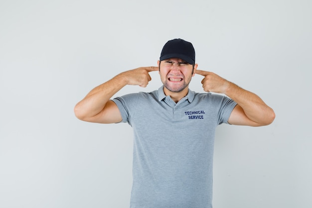 Young technician plugging ears with fingers in grey uniform and looking annoyed.