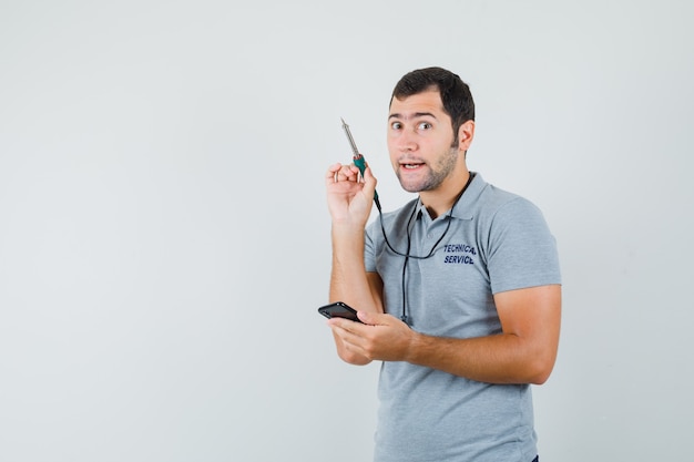 Young technician trying to open his smartphone by using drill  in grey uniform and looking focused.