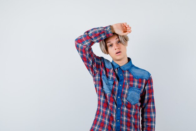 Young teen boy in checked shirt keeping hand on head and looking distressed , front view.