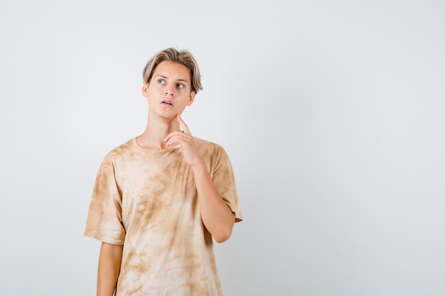 Free photo young teen boy keeping finger on jaw, looking up in t-shirt and looking pensive , front view.