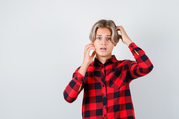 Young teen boy keeping hands on head in checked shirt and looking scared , front view.