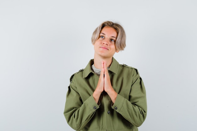 Free photo young teen boy keeping hands in praying gesture, looking up in green jacket and looking dreamy , front view.