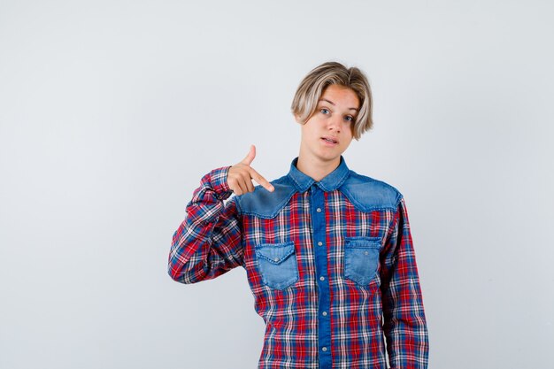 Young teen boy pointing down in checked shirt and looking hesitative. front view.