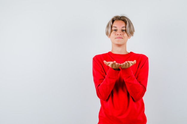 Free photo young teen boy in red sweater making giving or receiving gesture and looking cheerful , front view.