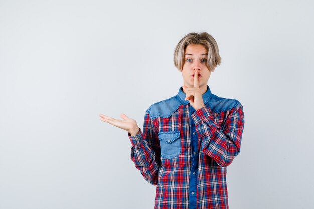 Young teen boy showing silence gesture, spreading palm aside in checked shirt and looking anxious , front view.