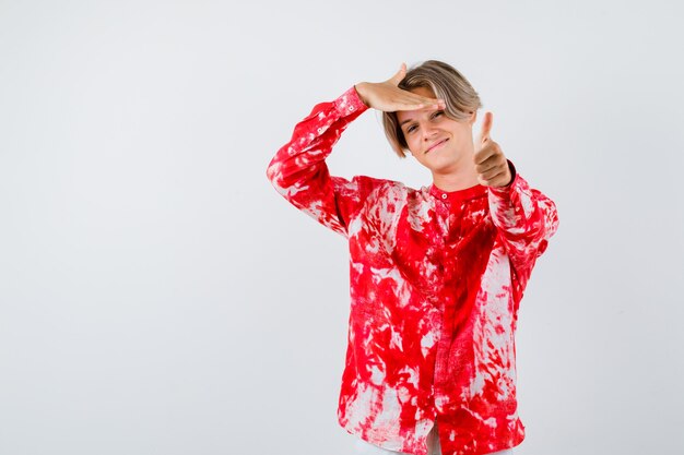 Young teen boy showing thumb up, keeping hand over head in shirt and looking merry , front view.