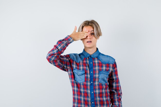 Young teen boy with hand on eye in checked shirt and looking wondered. front view.