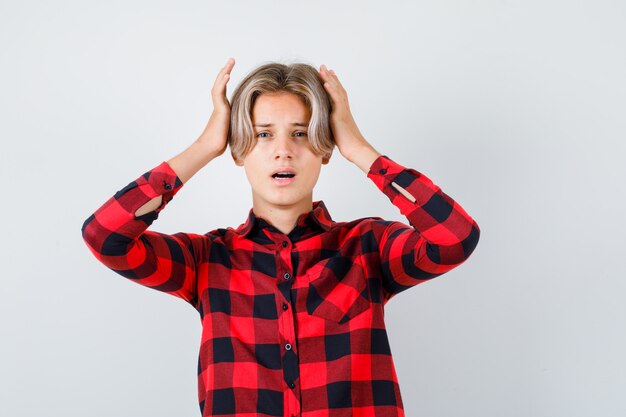 Young teen boy with hands on head in checked shirt and looking astonished. front view.