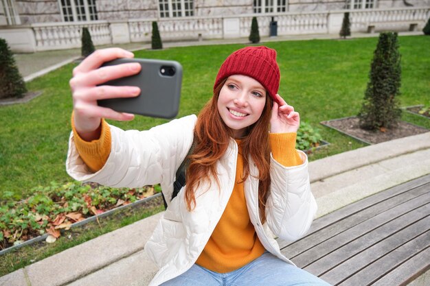 Young teen redhead girl sits on bench in park and takes selfie makes a photo of herself with smartph