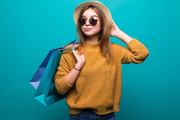 Young teen woman in sunglasses and hat holding shopping bags in her hands feeling so happiness isolated on green wall