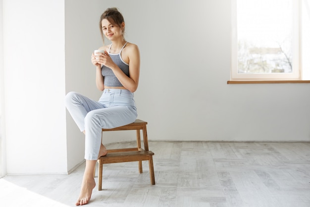 Young tender brunette woman smiling holding cup sitting on chair over white wall early in morning.