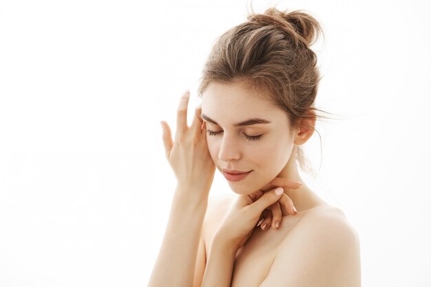 Young tender nude woman with bun posing over white background. Closed eyes.