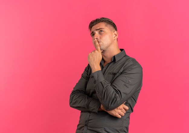 Young thoughtful blonde handsome man puts hand on chin looks up isolated on pink background with copy space