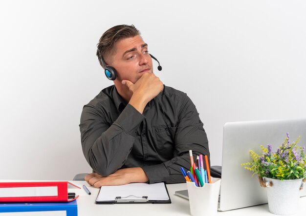 Young thoughtful blonde office worker man on headphones sits at desk with office tools using laptop puts hand on chin looking at side