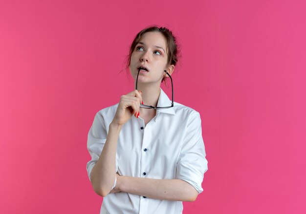 Young thoughtful blonde russian girl holds optical glasses close to mouth looking up isolated on pink space with copy space