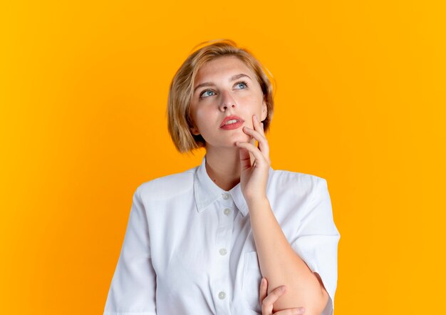 Young thoughtful blonde russian girl puts hand on face looking up isolated on orange background with copy space