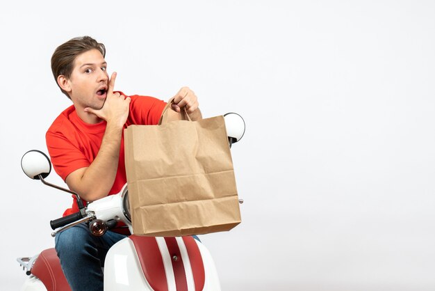 Young thoughtful courier guy in red uniform sitting on scooter holding paper bag on white wall