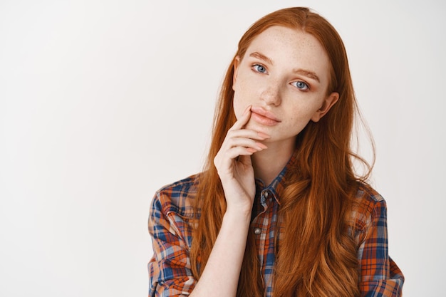 Free photo young thoughtful ginger girl in shirt, without makeup, looking at front and thinking, making choice, standing over white wall