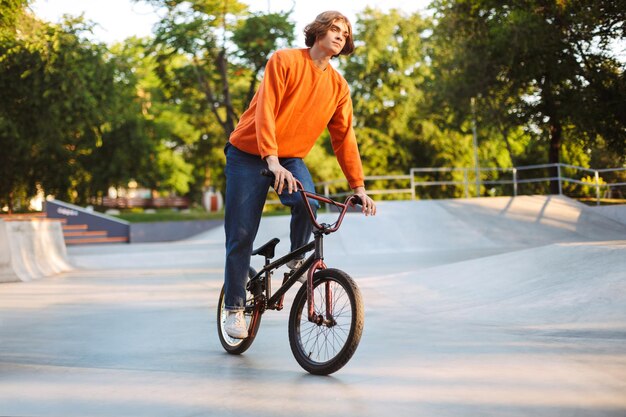 Young thoughtful guy in orange pullover and jeans dreamily riding bicycle at modern skatepark