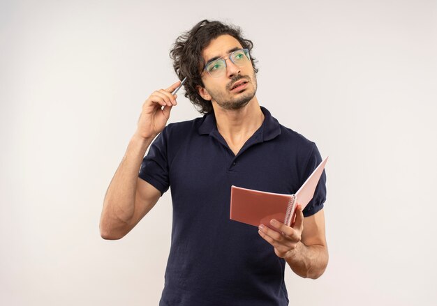 Young thoughtful man in black shirt with optical glasses holds notebook and puts pen on head isolated on white wall