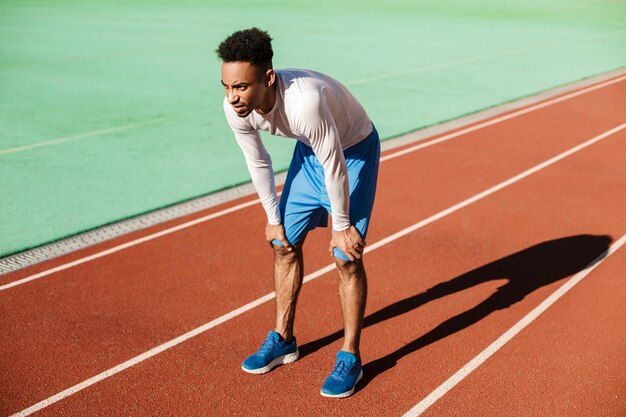 Young tired African American sportsman resting on racetrack after run at city stadium