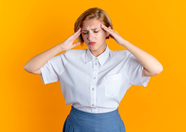 Free photo young tired blonde russian girl holds head isolated on orange background with copy space