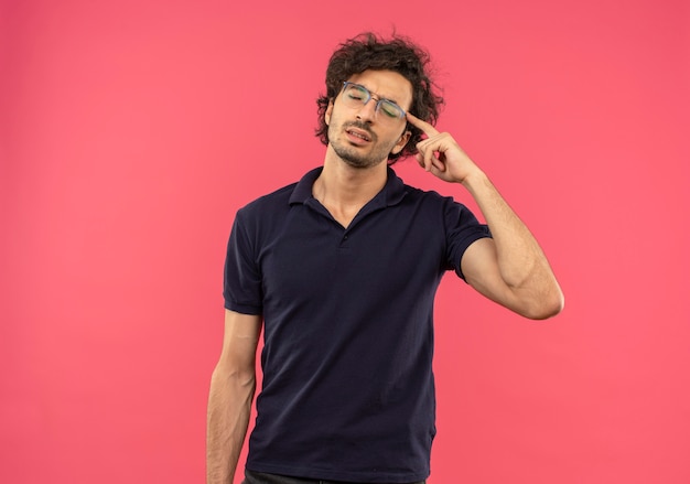 Young tired man in black shirt with optical glasses puts finger on head isolated on pink wall