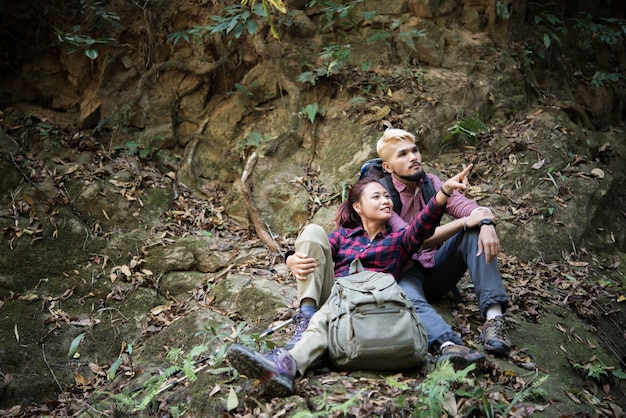 Free photo young tourist couple take a rest near waterfall while hiking in forest looking away in search of attractions. travel concept.